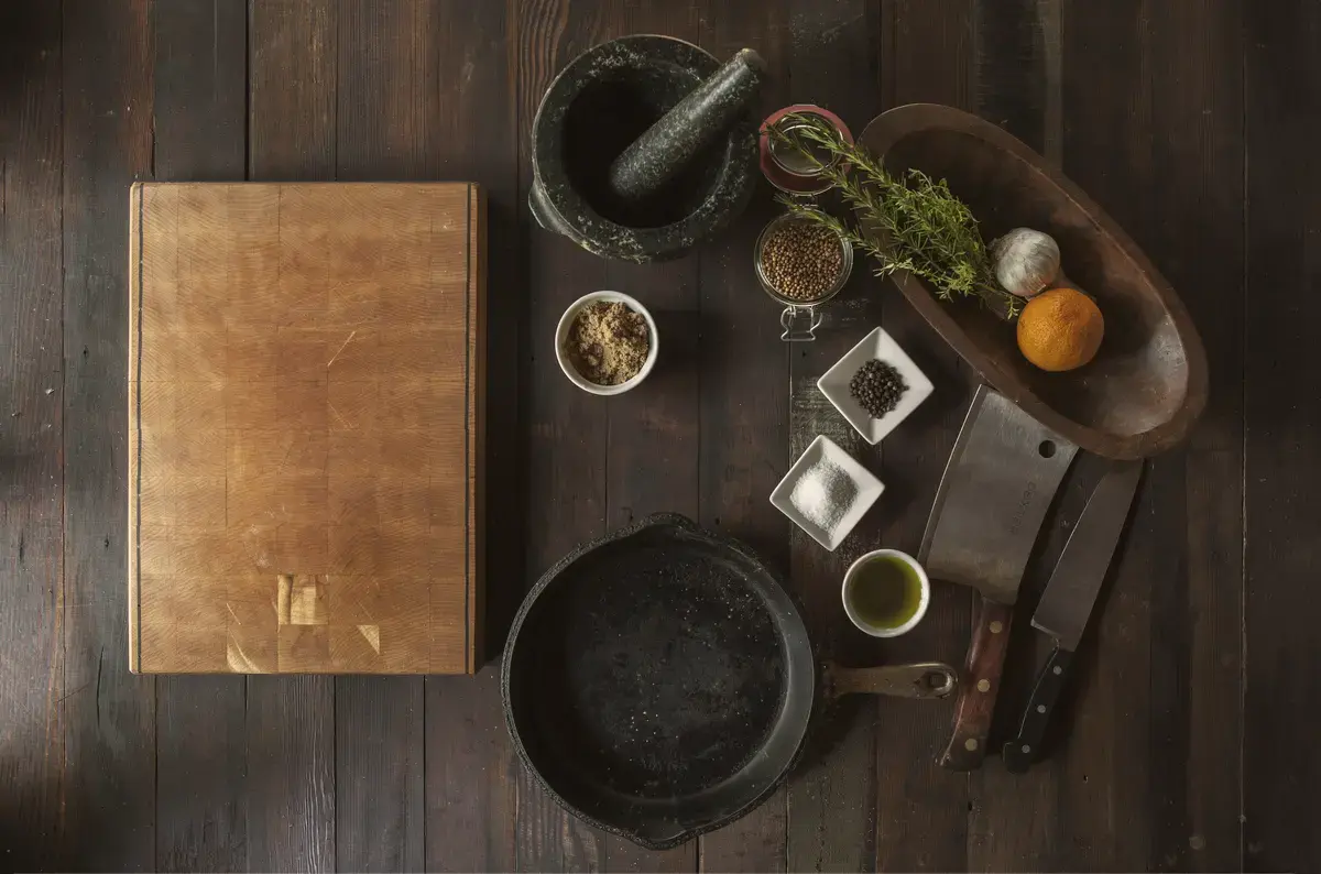 Overhead view of a cook's table: chopping board, cast-iron pan, knives, mortar and pestle, herbs and spices on dark wood