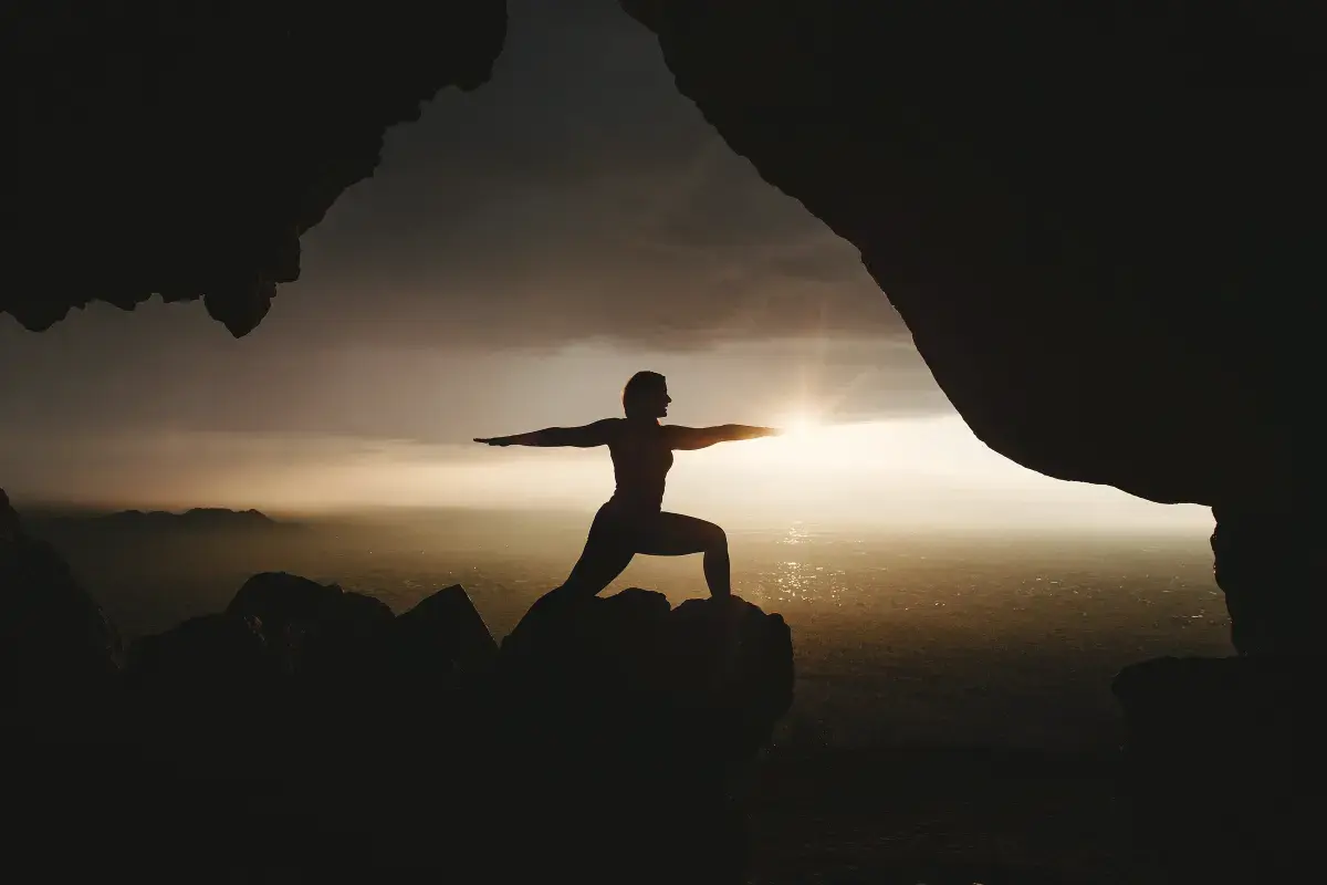 Silhouette in a yoga warrior pose at sunrise, framed by a rocky cave opening